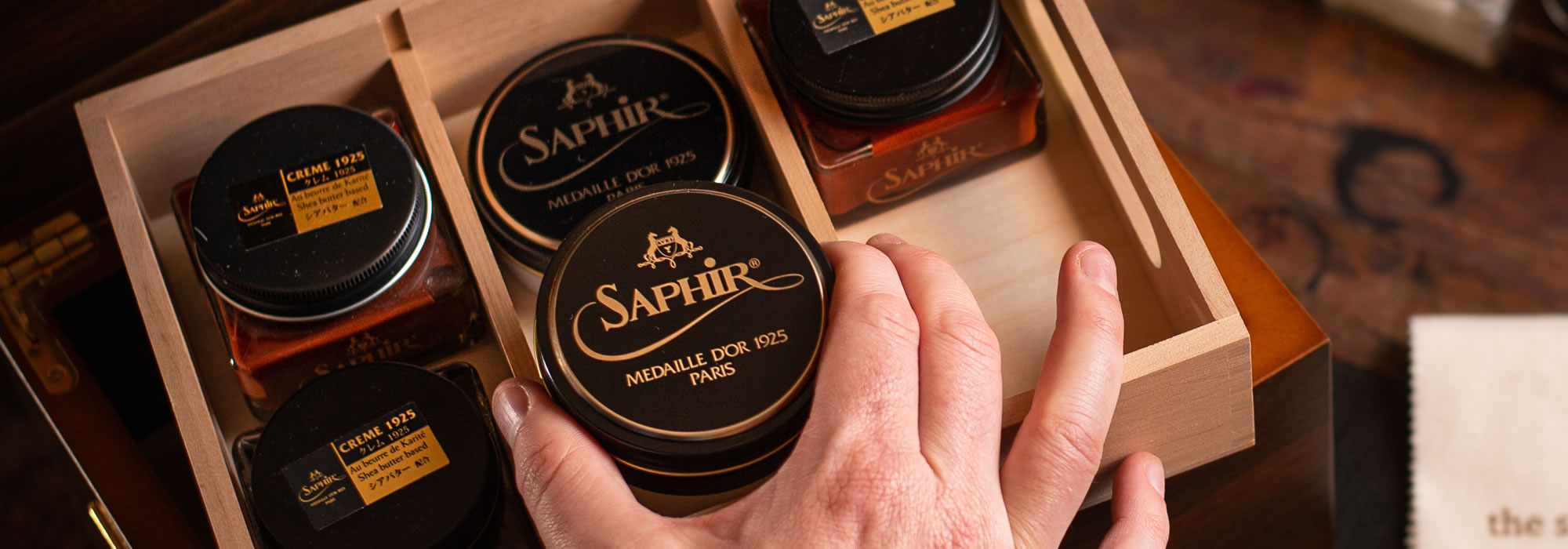 A hand reaches into a wooden box filled with Saphir shoe care products, conveying a sense of luxury and meticulous shoe maintenance.