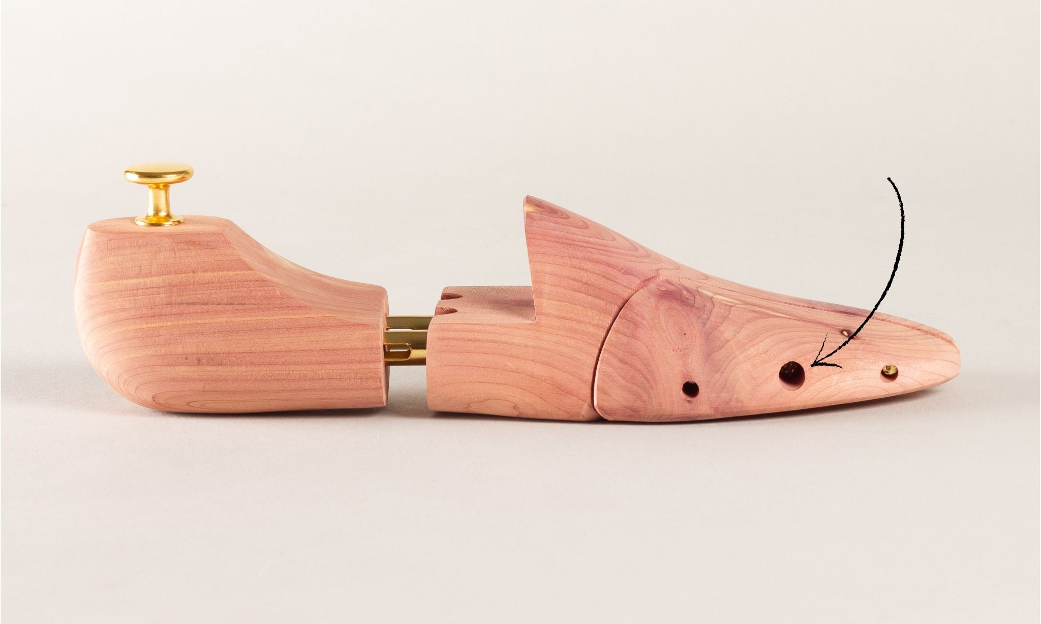 A cedar wood shoe tree lies on its side, featuring a brass knob and joints. A black arrow points to a hole on the right.