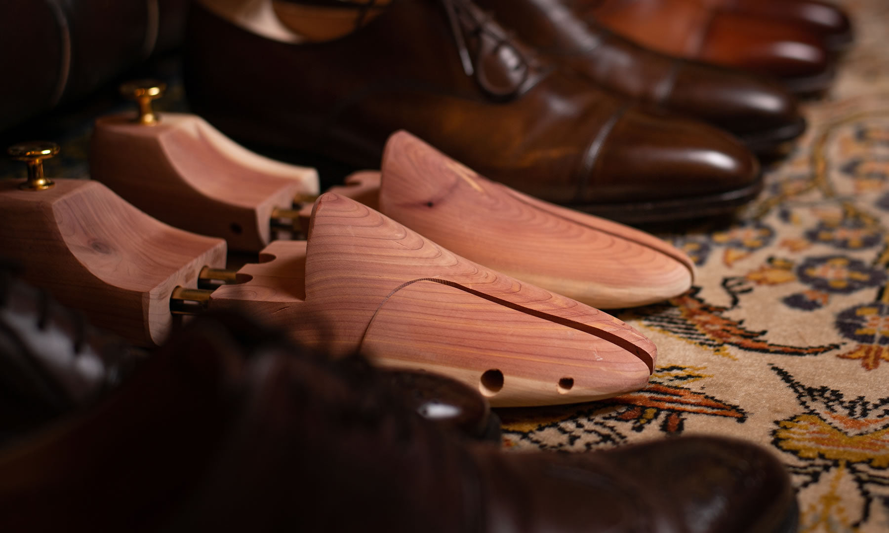 A pair of wooden shoe trees placed on a patterned rug, with several pairs of polished brown leather shoes in the background.