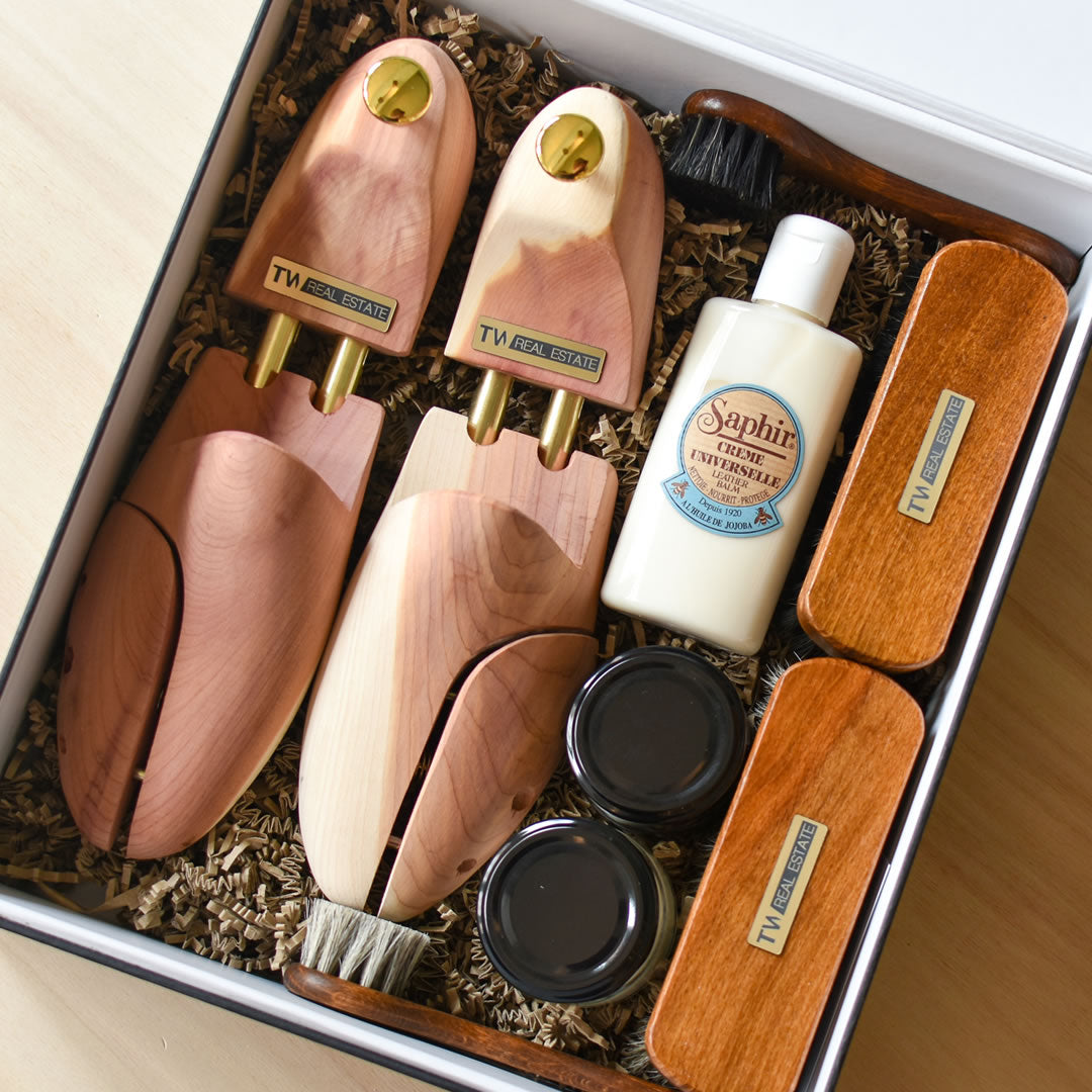 A shoe care kit in a box, including two wooden shoe trees, a bottle of Saphir cleaner, two brushes, two wooden containers, two small jars, and packing material surrounding the items.